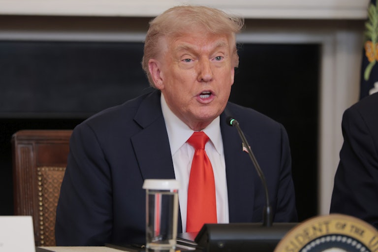 Donald Trump, hunched over at a table, speaks into a mic.