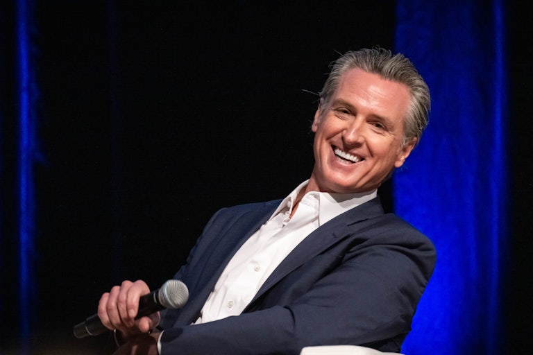 Gavin Newsom holds a mic in his hand and laughs as he leans back in his chair.