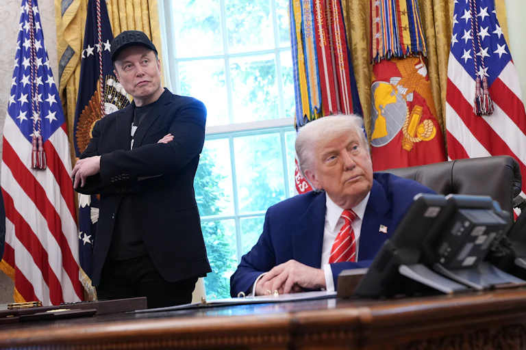 Elon Musk, wearing a black DOGE cap, stands and crosses his arms while Donald Trump sits at this desk in the Oval Office of the White House.