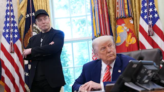 Elon Musk, wearing a black DOGE cap, stands and crosses his arms while Donald Trump sits at this desk in the Oval Office of the White House.