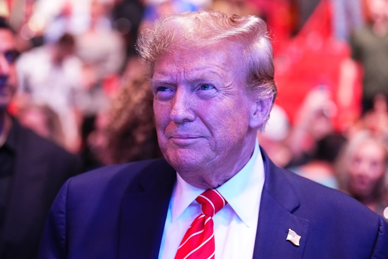 Donald Trump smiles at something off camera. He is wearing a suit with an American flag lapel pin. There is a crowd (unfocused) in the background.