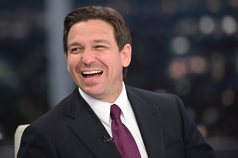 Ron DeSantis, wearing a suit and tie, laughs and looks off camera