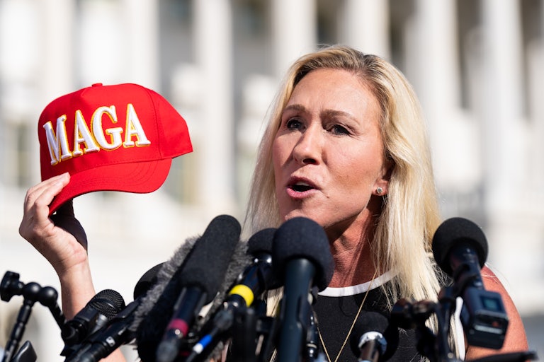 Marjorie Taylor Greene holds a red MAGA cap and speaks before a dozen of mics. She is presumably outside the Capitol.