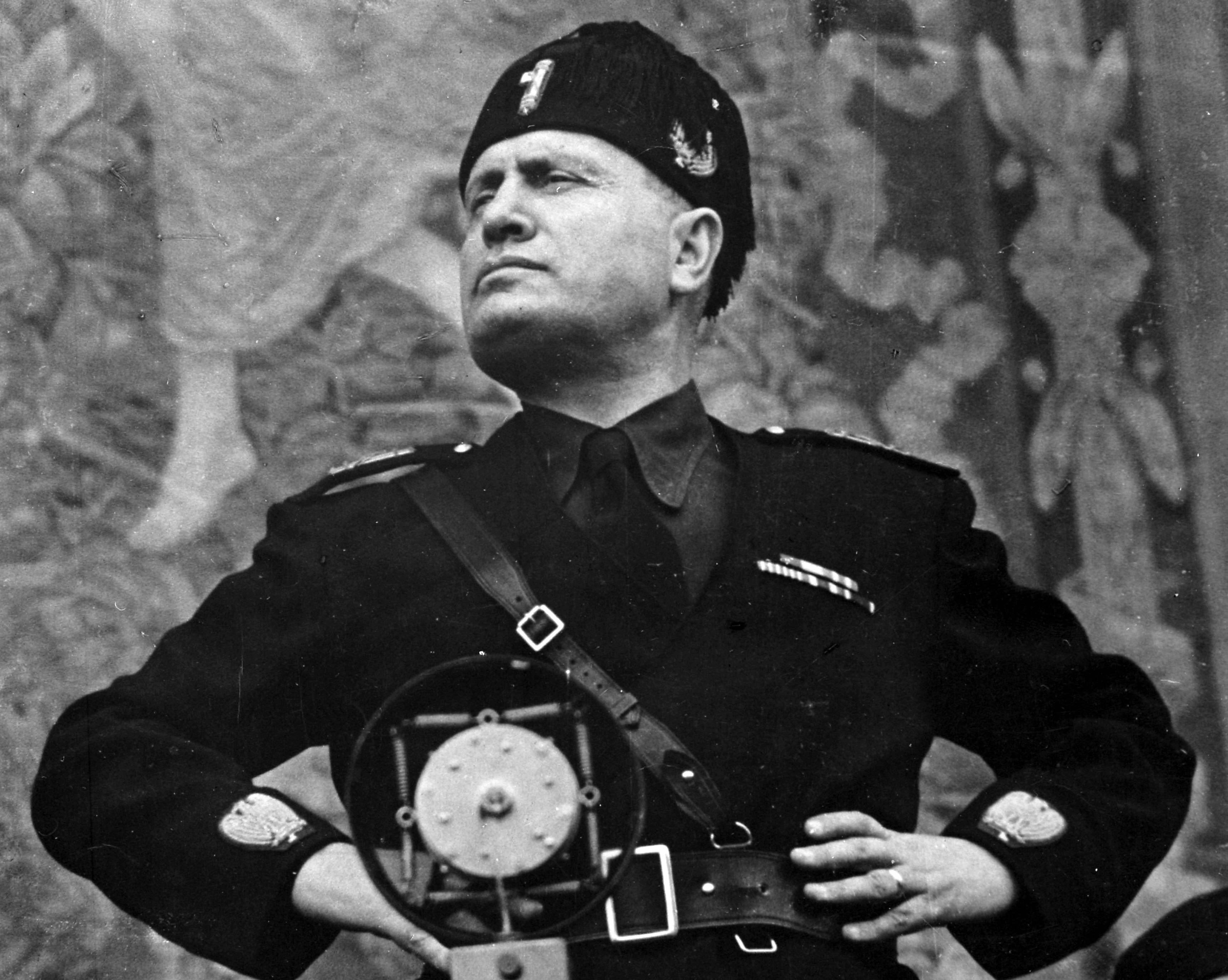 Italian fascist dictator Benito Mussolini stands with his hands on his hips as he stares defiantly into the middle distance.