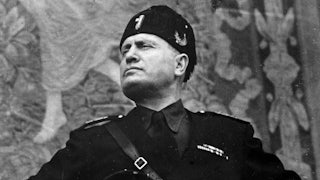 Italian fascist dictator Benito Mussolini stands with his hands on his hips as he stares defiantly into the middle distance.