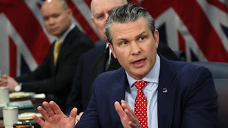 Pete Hegseth speaks at a table and raises both hands as if in defense.