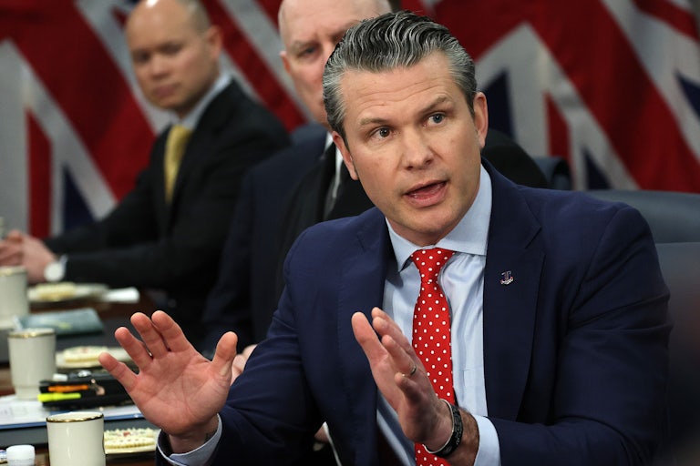 Pete Hegseth speaks at a table and raises both hands as if in defense.