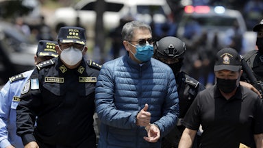 Juan Orlando Hernández, in handcuffs and flanked by police, gives a thumbs up.