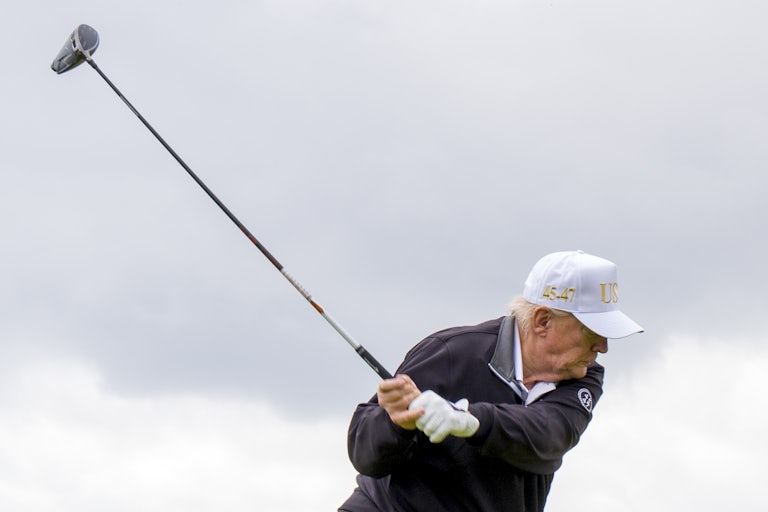 Donald Trump swings a golf club and wears a white USA cap.
