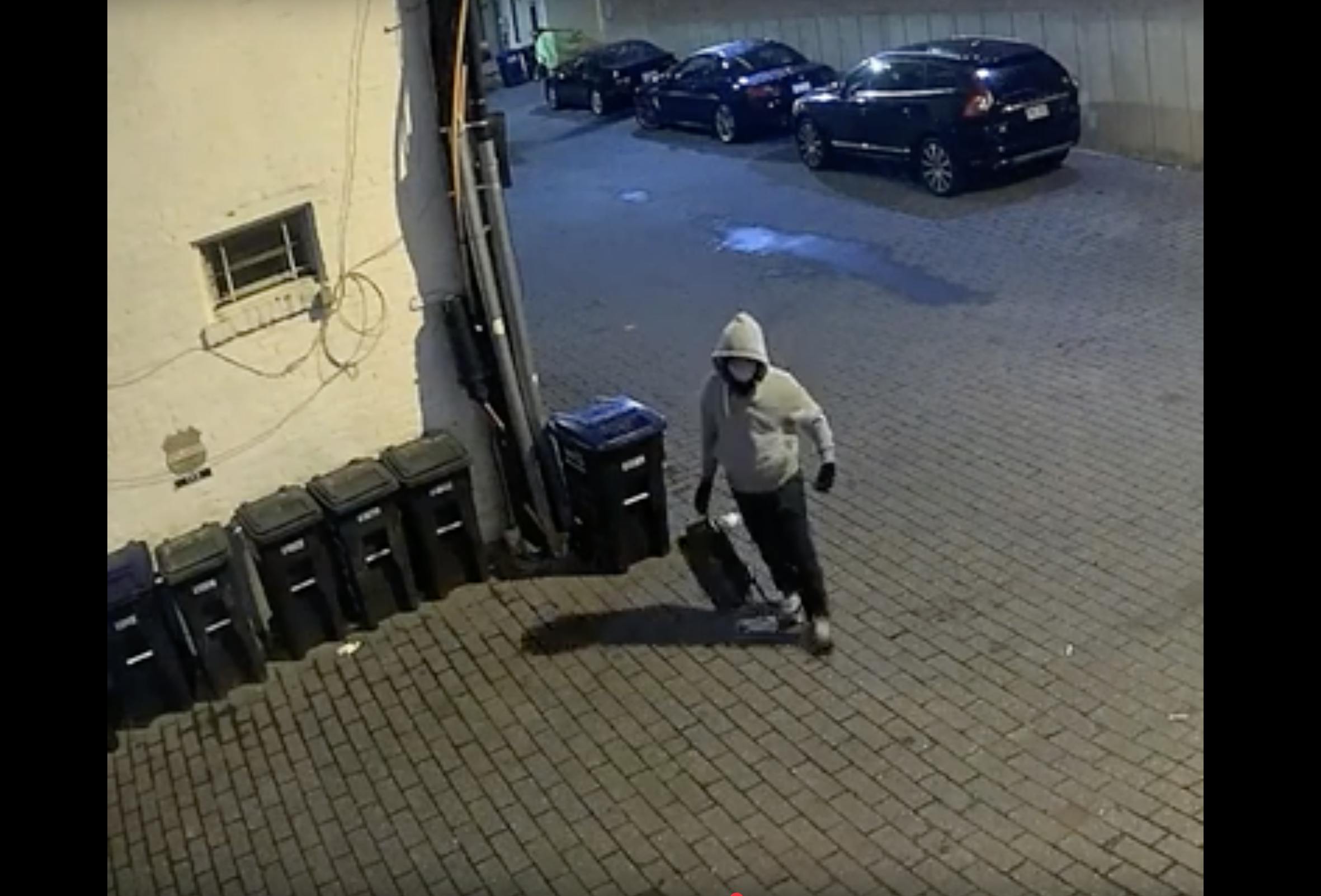 The pipe bombing suspect wears a grey hoodie, a face mask, and carries a backpack outside near a row of trashcans.