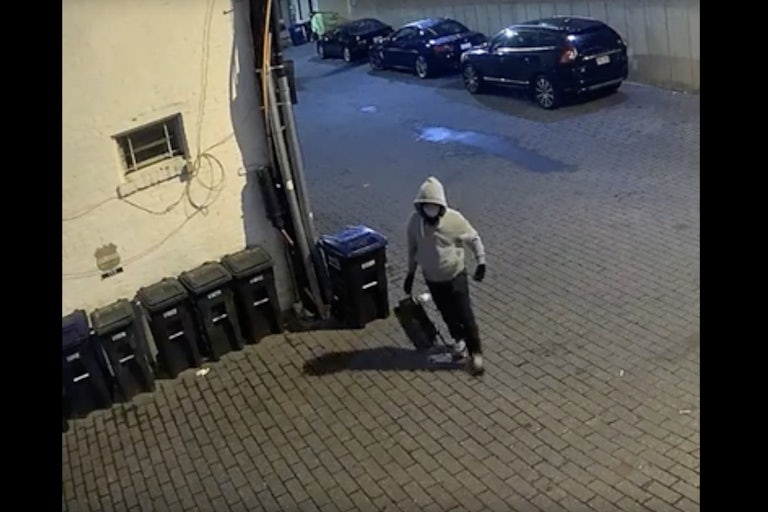 The pipe bombing suspect wears a grey hoodie, a face mask, and carries a backpack outside near a row of trashcans.