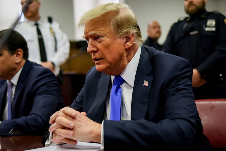 Donald Trump grimaces in court, hands clasped on the table. His lawyer sits beside him and in the background are two security guards.