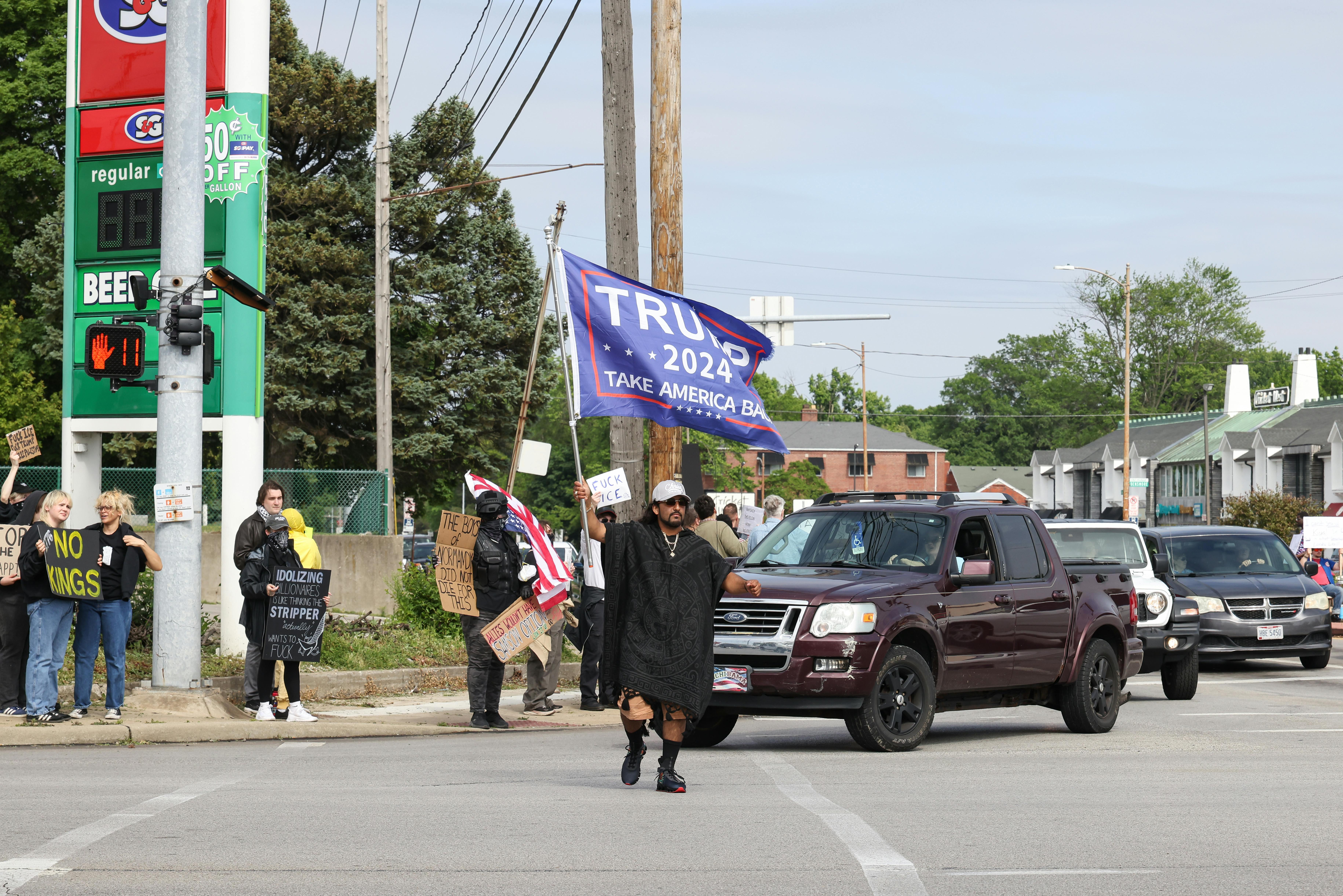 No Kings protesters stand on the sidewalk a street corner while one person crossing the road waves a Trump 2024 flag.