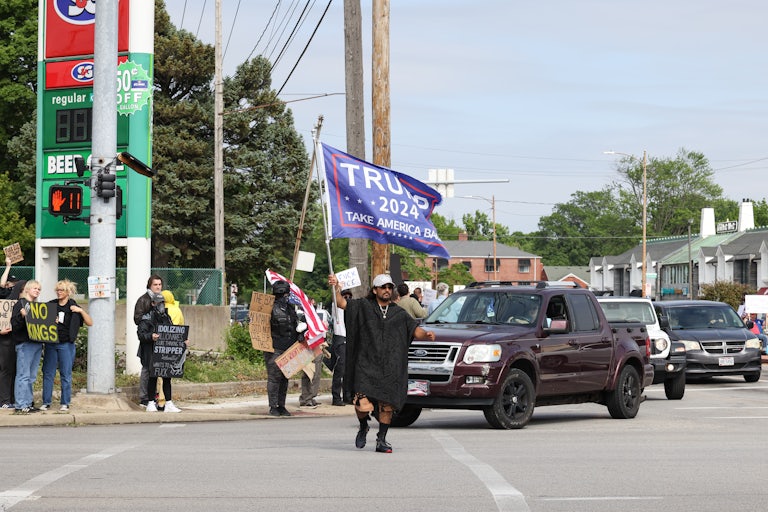 No Kings protesters stand on the sidewalk a street corner while one person crossing the road waves a Trump 2024 flag.