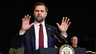 Vice President JD Vance speaks at a podium and raises both hands as if in defense