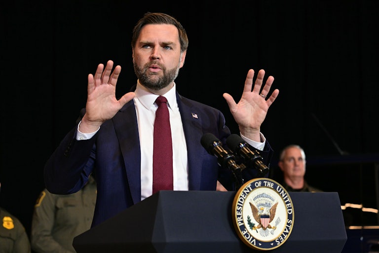 Vice President JD Vance speaks at a podium and raises both hands as if in defense
