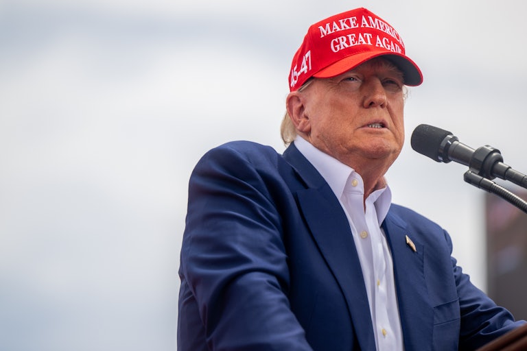 Trump is wearing a red Make America Great again cap and speaking at a mic