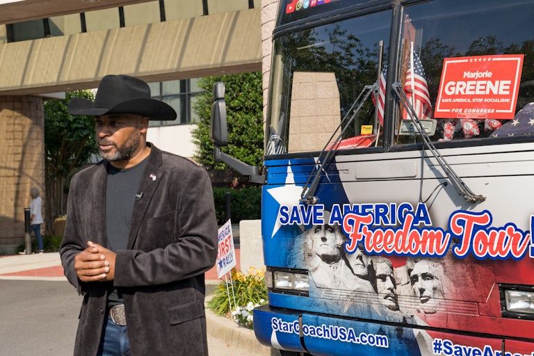 A cowboy hat-clad Marcus Flowers stands in front of Marjorie Taylor Greene's bus speaking to reporters.