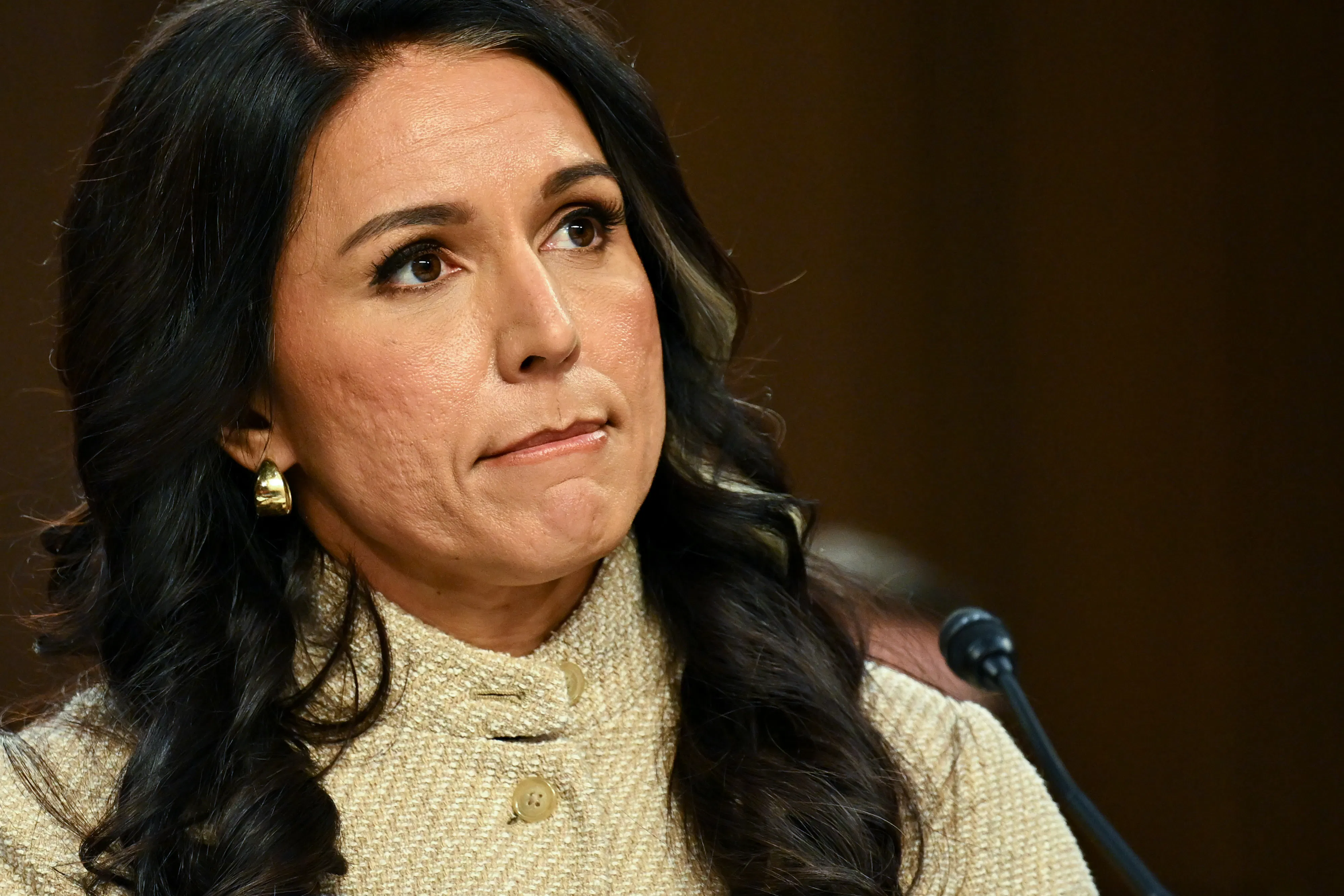 Tulsi Gabbard, wearing a beige cardigan, sits with her lips pursed before a dark backdrop. A microphone is visible in front of her.