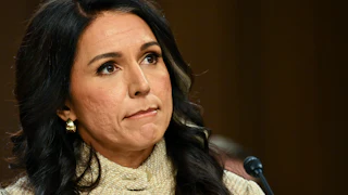 Tulsi Gabbard, wearing a beige cardigan, sits with her lips pursed before a dark backdrop. A microphone is visible in front of her.