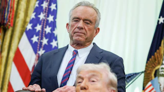 Robert F. Kennedy Jr. stands with his hands on the back of Donald Trump's desk chair in the Oval Office. Trump sits in the chair and makes a pouty face.