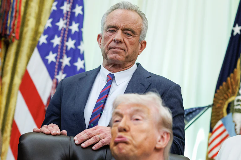 Robert F. Kennedy Jr. stands with his hands on the back of Donald Trump's desk chair in the Oval Office. Trump sits in the chair and makes a pouty face