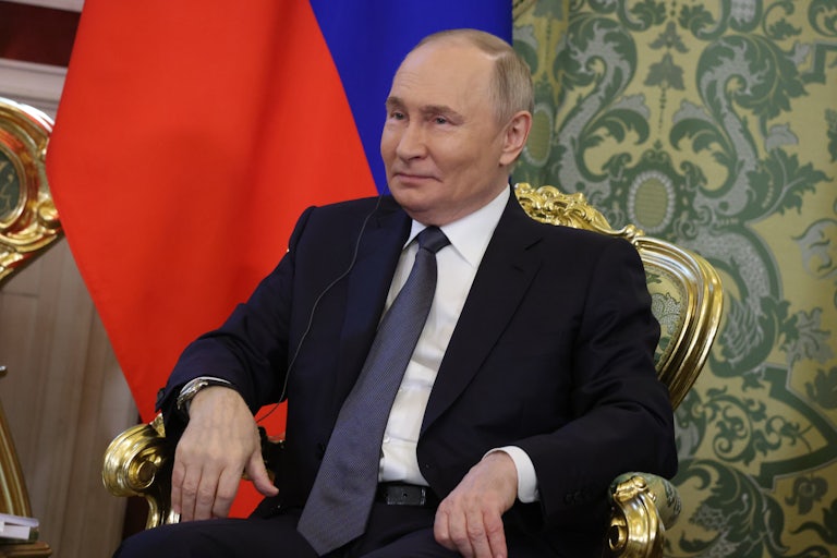 Russian President Vladimir Putin smiles while sitting on a golden chair in front of a Russian flag.