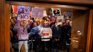 Demonstrators protest behind a glass holding signs like "Hoosiers Play Fair No Redistricting" and "Democracy Doesn't Get Redrawn."