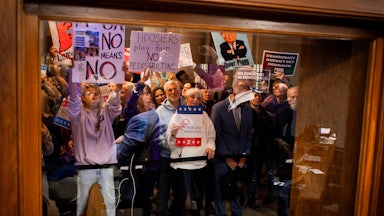 Demonstrators protest behind a glass holding signs like "Hoosiers Play Fair No Redistricting" and "Democracy Doesn't Get Redrawn."