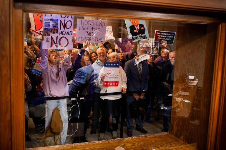 Demonstrators protest behind a glass holding signs like "Hoosiers Play Fair No Redistricting" and "Democracy Doesn't Get Redrawn."