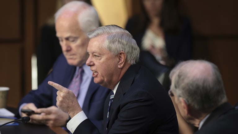 Senator Lindsey Graham speaks in the foreground while pointing a finger. Senator John Cornyn is seated beside him in the background.