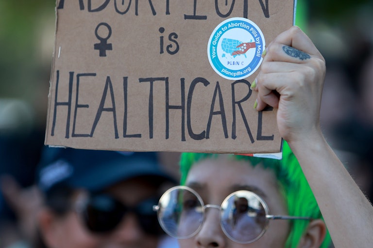 Someone with green hair and sunglasses holds a cardboard sign reading "Abortion is healthcare."