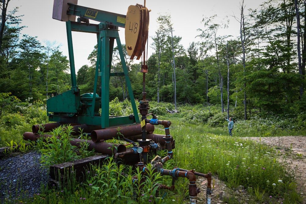 A pumpjack stands in a forest clearing, with a person in the background.