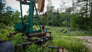 A pumpjack stands in a forest clearing, with a person in the background.