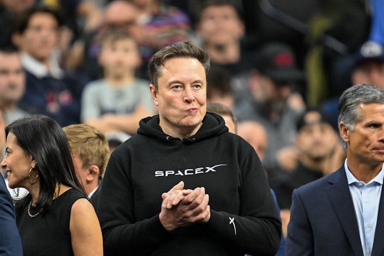 Elon Musk frowns and stands with his hands clasped while attending a college wrestling championship in Philadelphia