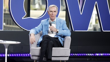 Bill Nye the Science Guys sits on a chair on stage and speaks with a mic in his hand.