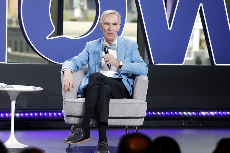 Bill Nye the Science Guys sits on a chair on stage and speaks with a mic in his hand.