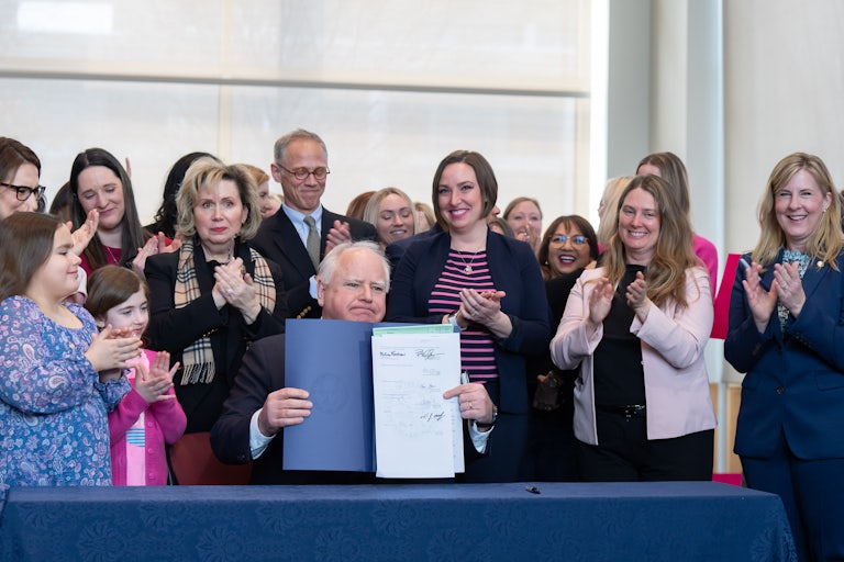Tim Walz sits at a desk and holds a signed copy of the bill up for the cameras. About two dozen people (mostly women) cheer around him.