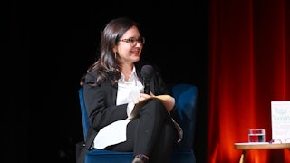 Bari Weiss crosses her legs as she sits on a chair on stage, talkign to someone else (unpictured).