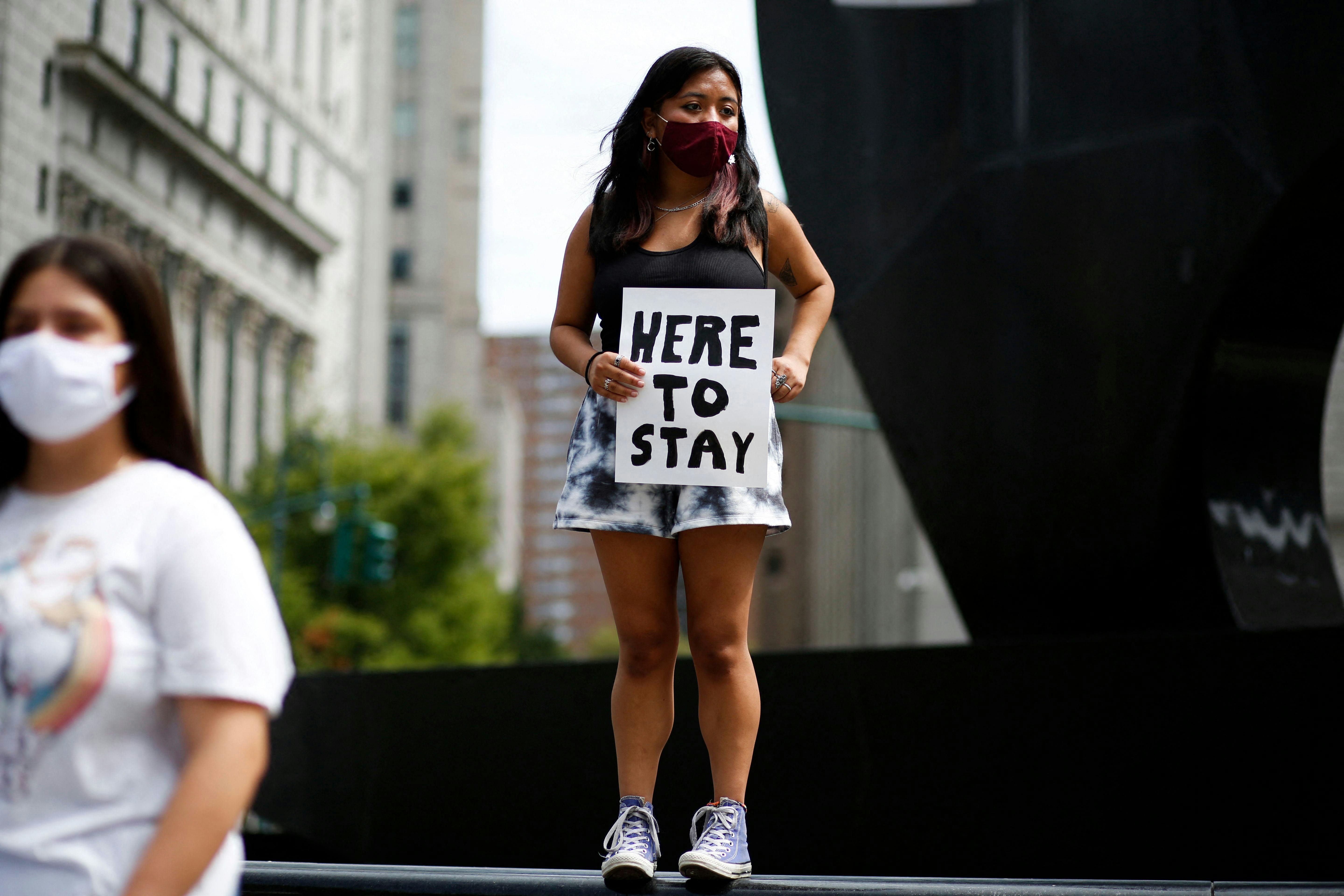 A young woman holds a banner reading “Here to Stay”