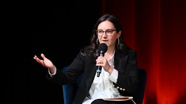 Bari Weiss gestures while speaking into a microphone while seated.