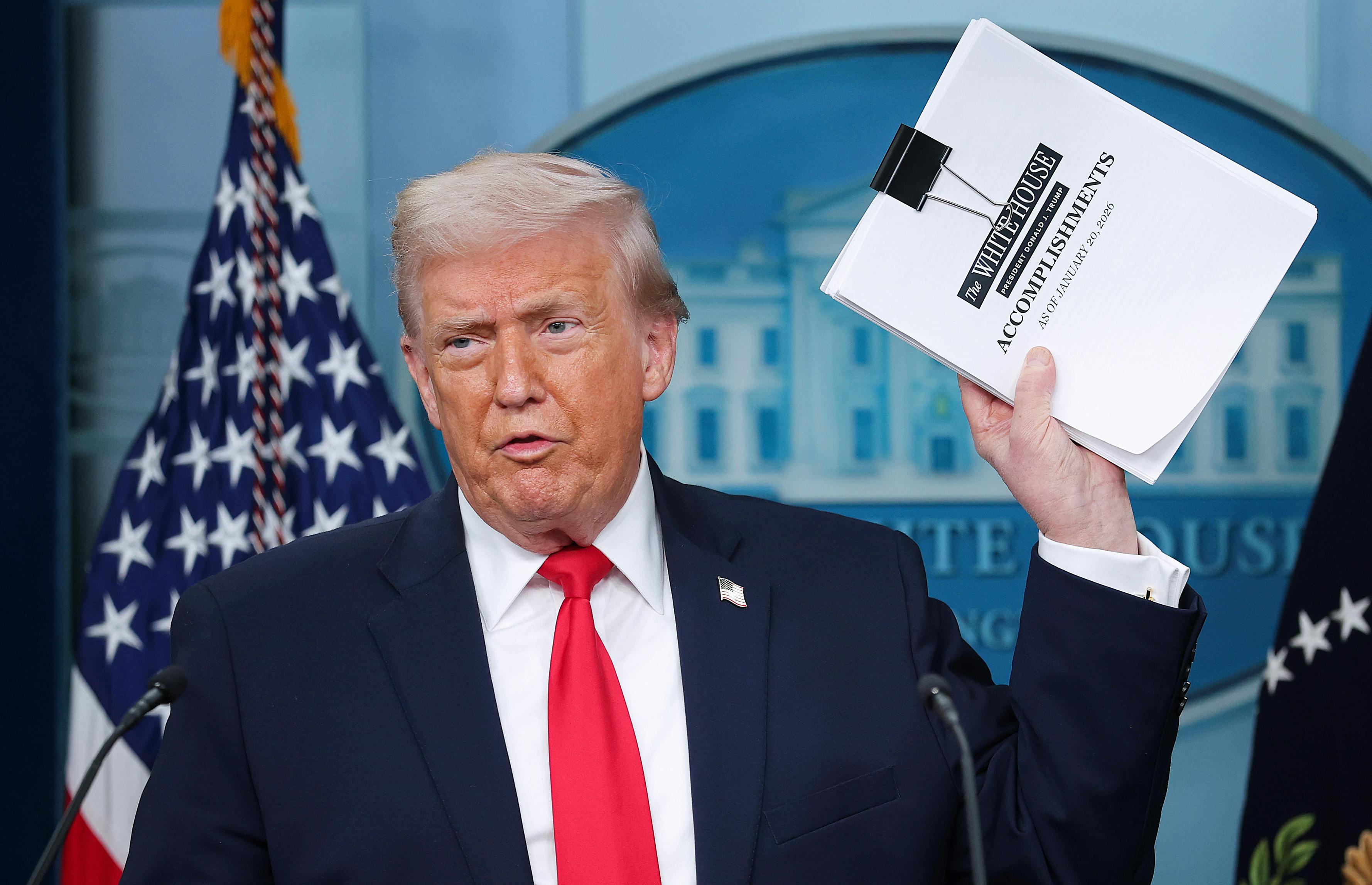Donald Trump speaks in the White House press briefing room and holds up a stack of papers that reads "White House accomplishments."
