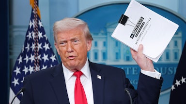 Donald Trump speaks in the White House press briefing room and holds up a stack of papers that reads "White House accomplishments."