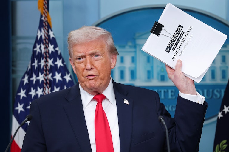 Donald Trump speaks in the White House press briefing room and holds up a stack of papers that reads "White House accomplishments."