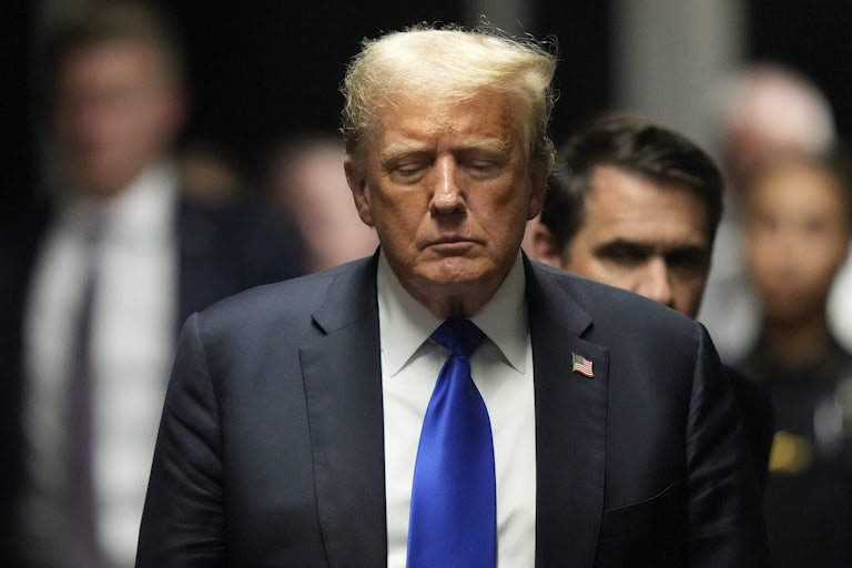 A man, wearing a black suit and blue tie, looks down while walking. he is sad because he got in trouble for doing crimes