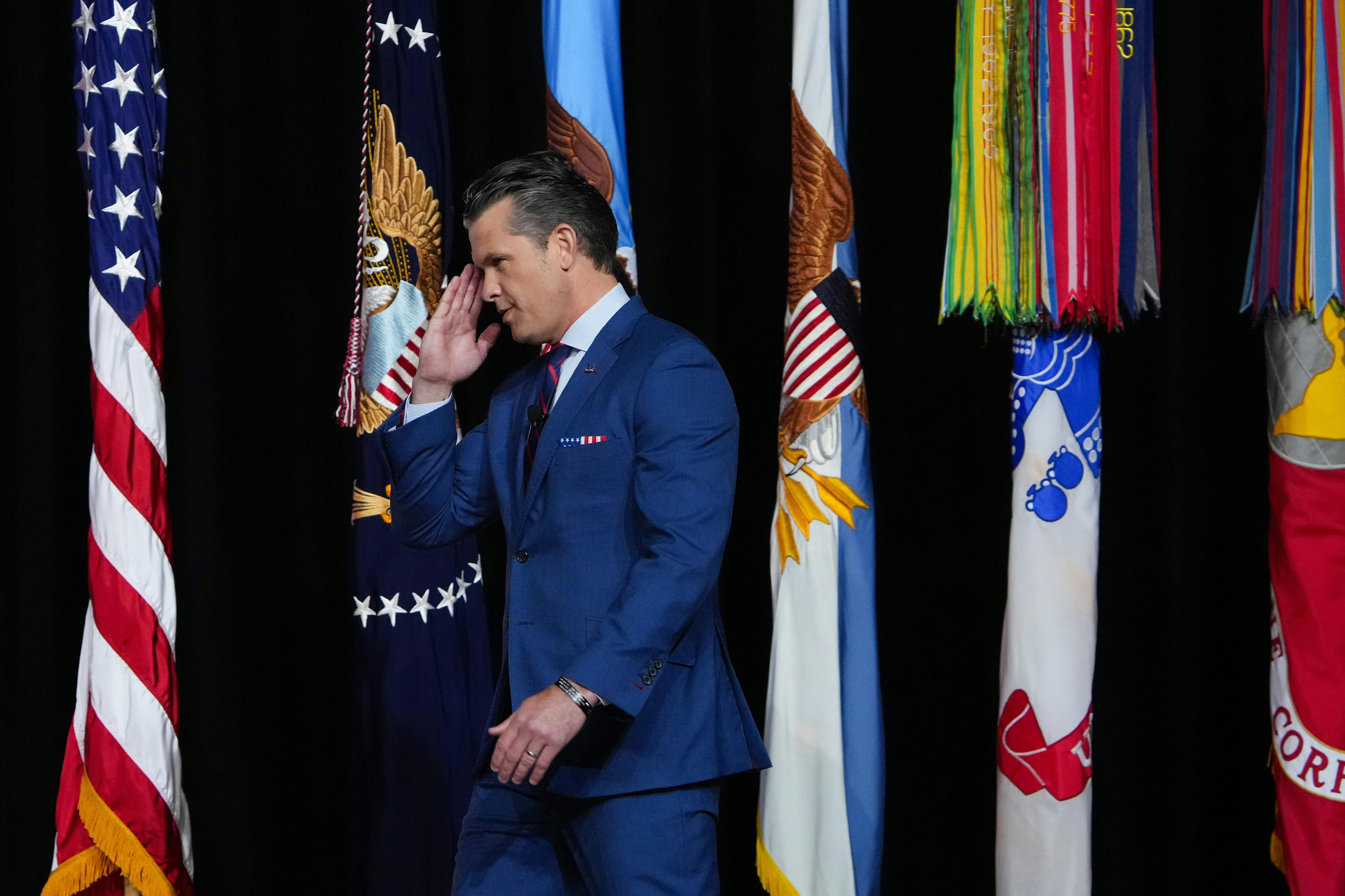 Defense Secretary Pete Hegseth salutes s he walks in front of several flags.