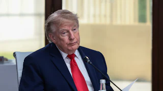 A fatigued Donald Trump, sitting down wearing a blue suit and red tie.