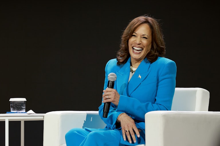 Kamala Harris laughing as she's seated on a white armchair. She's wearing a bright blue pantsuit and holding a mic in her hand.
