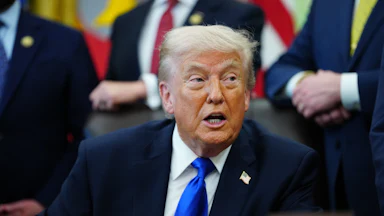 Donald Trump turns to the side and speaks while sitting at his desk in the Oval Office