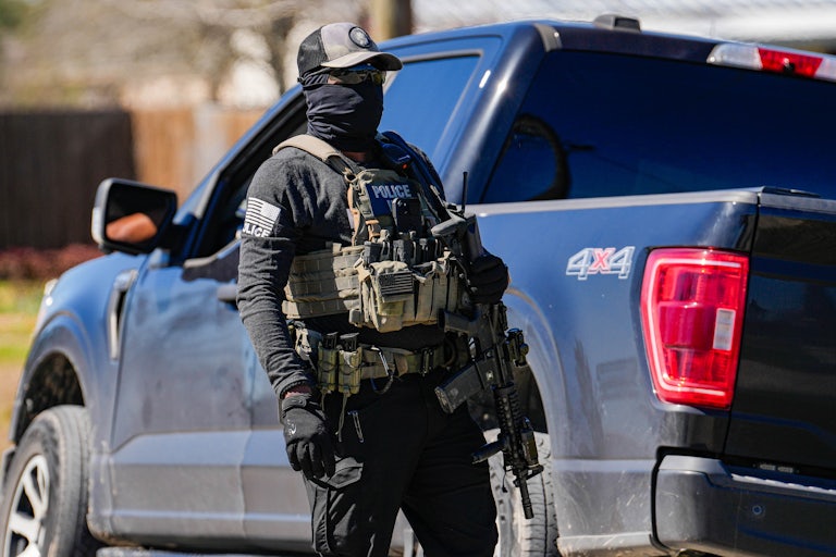 An armed agent carries a massive gun and other weapons. His face is completely covered with a cap, a balaclava, and sunglasses.